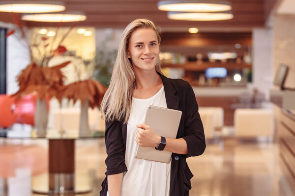A smiling front desk agent with a tablet in hand greets guests in the lobby.