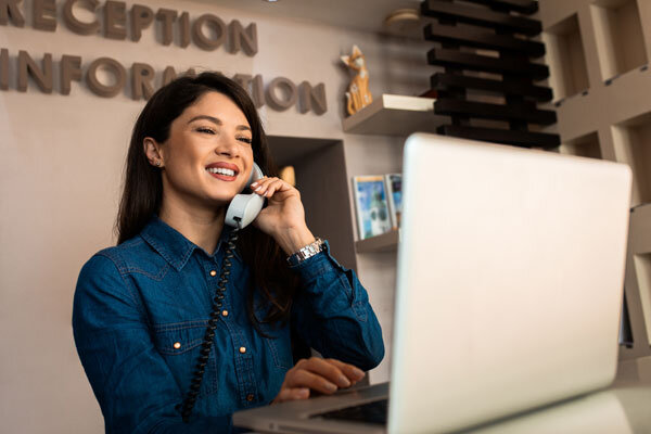 A smiling hotel receptionist helps a potential guest on the phone while confidently referring to the property management system.