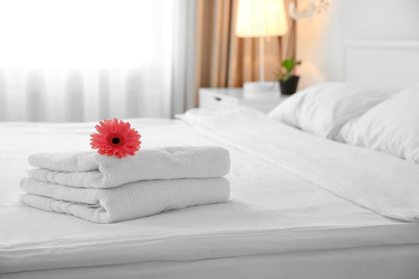 A freshly made hotel bed with clean, white linens and carefully folded white towels adorned with a pink flower welcomes guests.