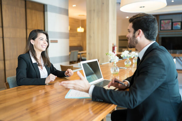 A hotel manager interviews a smiling front desk candidate in a hotel meeting room.