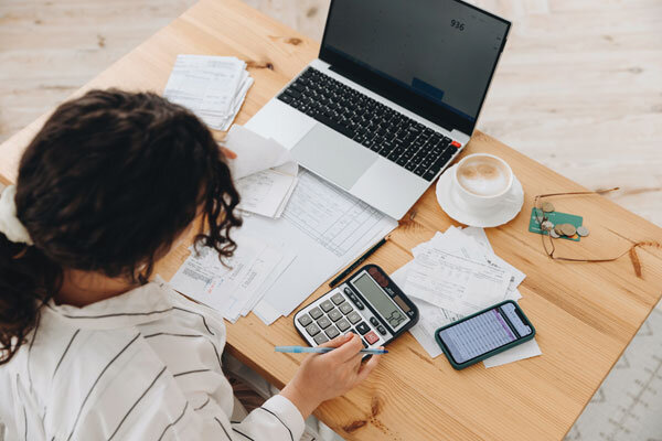 A woman sits at a desk with a laptop, receipts, calculator, and a cup of coffee, calculating expenses.