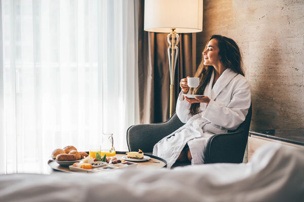 A smiling woman in a fluffy bathrobe enjoys breakfast in her hotel room.