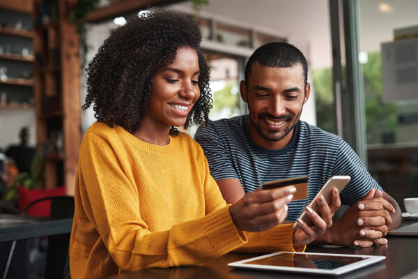 A smiling couple seated at a cafe makes a hotel booking online using a smartphone