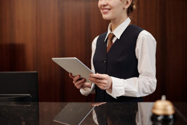 A smiling hotel receptionist with tablet device in hand greets guests at the front desk.