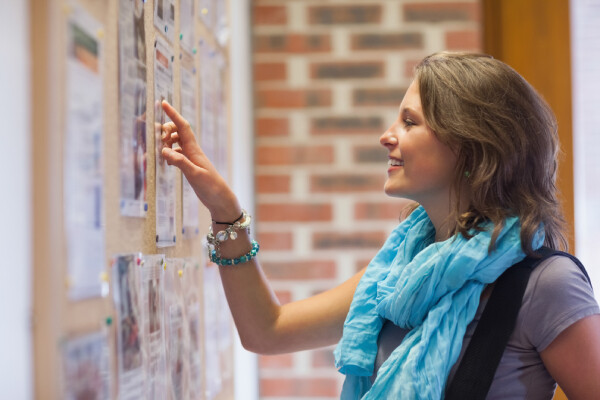 A young, smiling woman looks for job opportunities on a community noticeboard.