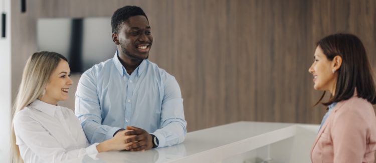 Happy smiling multiethnic couple standing at a modern reception desk, talking with a friendly female hotel receptionist